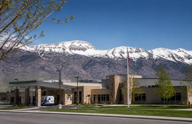 Modern stone-faced rural hospital with an ambulance under the entry canopy and snow-capped peaks behind — Beaver Valley Hospital is county-owned and contracts with carriers independently