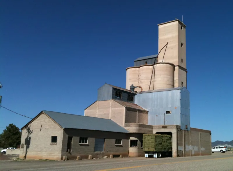 Historic concrete grain elevator and feed warehouse in Enterprise, Utah with a hay-hauling flatbed truck loaded with alfalfa bales out front — Escalante Valley farm-and-ranch country in ZIP 84725.