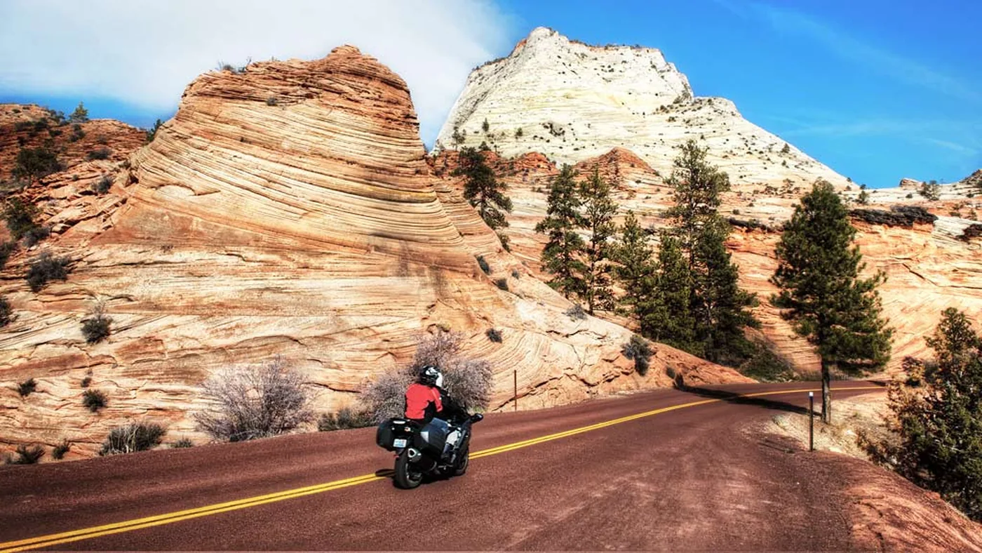 Motorcycle rider on the red asphalt of Zion–Mt. Carmel Highway with white Navajo sandstone domes above — OnPoint covers Southern Utah's national-park rides.