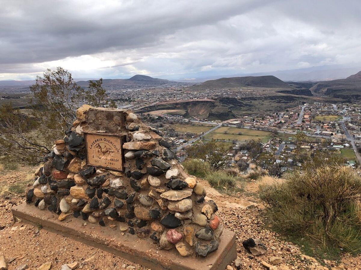 View from the La Verkin Overlook (settled 1891) with a rock cairn monument in the foreground looking down on the town, the Virgin River canyon, and the volcanic cinder cones above Hurricane Valley — the 84745 streets and SR-9 corridor that shape local auto insurance pricing