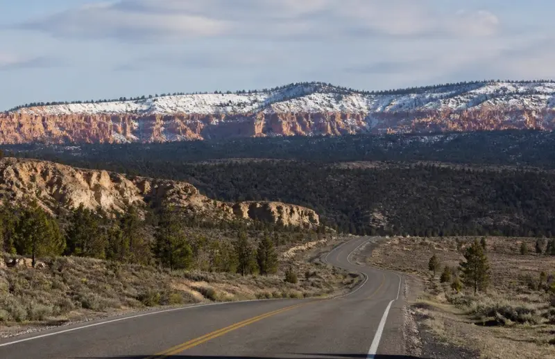 Empty two-lane highway curving through sagebrush and pines toward snow-topped orange cliffs near Bryce Canyon — winter driving distance shapes Garfield County health plan choices