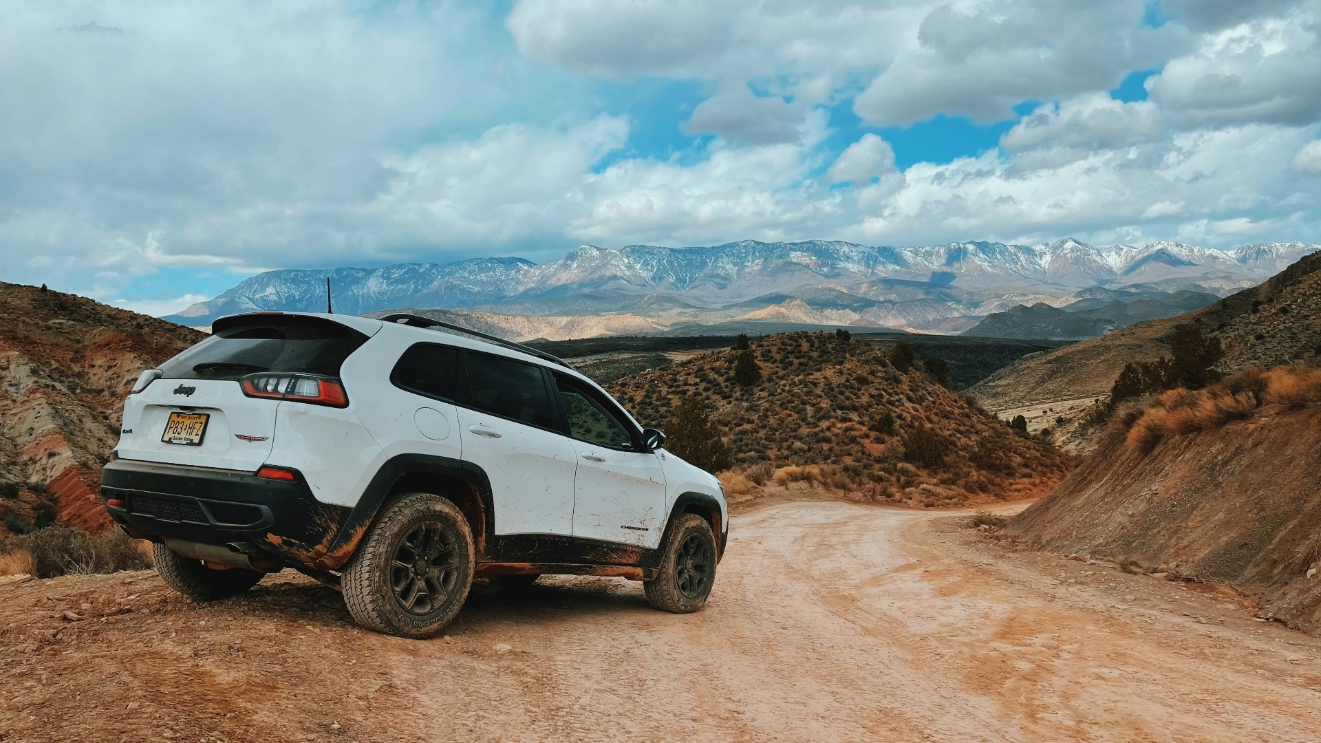 A muddy Jeep Cherokee Trailhawk parked on a backcountry dirt road outside La Verkin, Utah with snow-capped Pine Valley Mountains in the distance — off-road and farm-road exposure on La Verkin auto policies