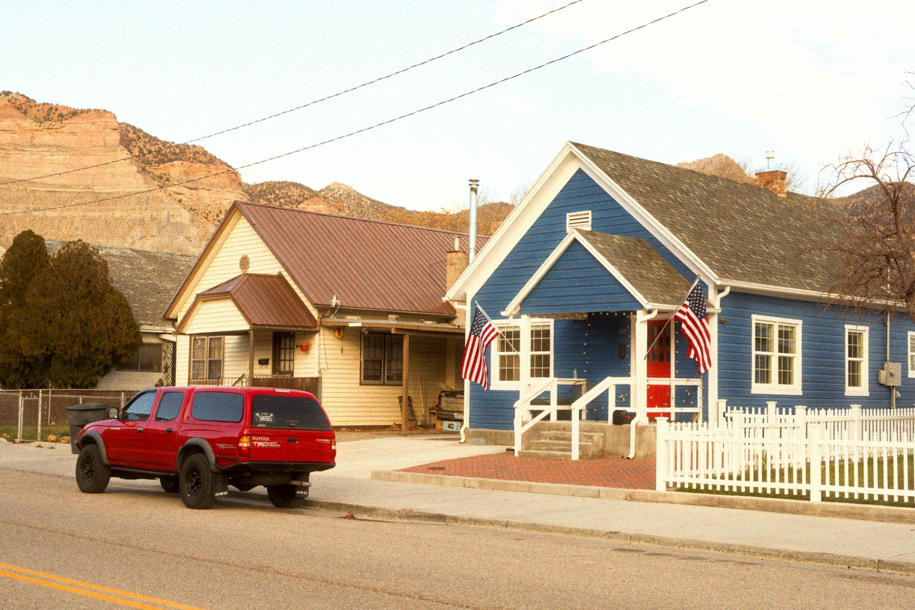 Small-town Enterprise, Utah homes with white picket fence and American flags along a quiet street, red sandstone cliffs rising behind the neighborhood — the downtown Enterprise streetscape and rebuild profile that shapes 84725 homeowners insurance