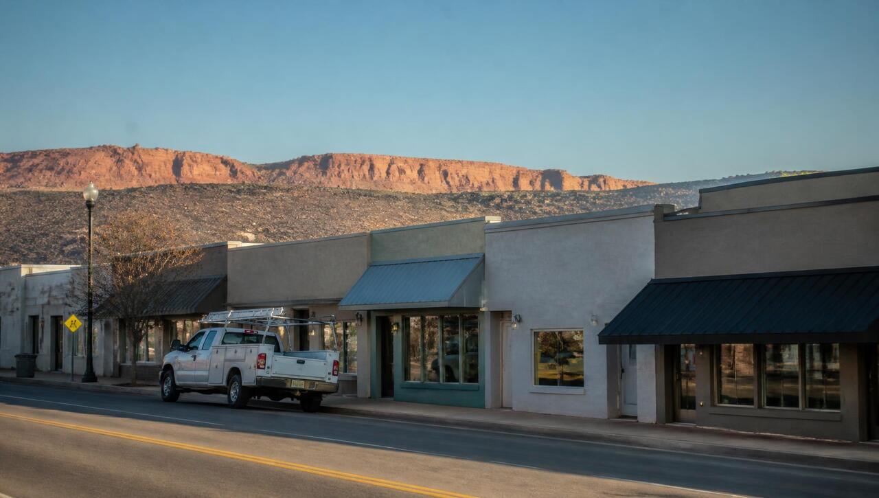 Main street storefronts with awnings and a work van at the curb in Washington, Utah, red rock ridges behind (ZIP 84780) — business insurance, BOP, general liability, commercial property and workers' comp for Washington, Utah employers