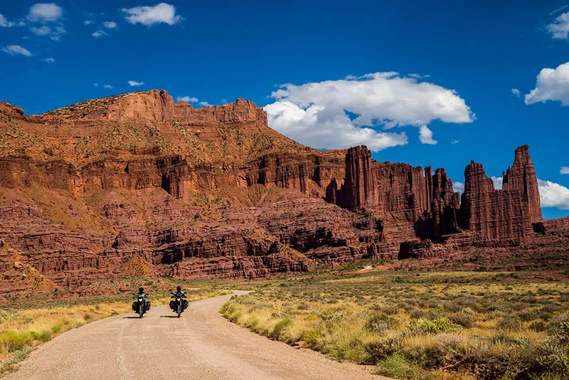 Two adventure motorcyclists riding a dirt road toward red-rock Fisher Towers in Moab, Utah — OnPoint Utah Motorcycle Insurance.